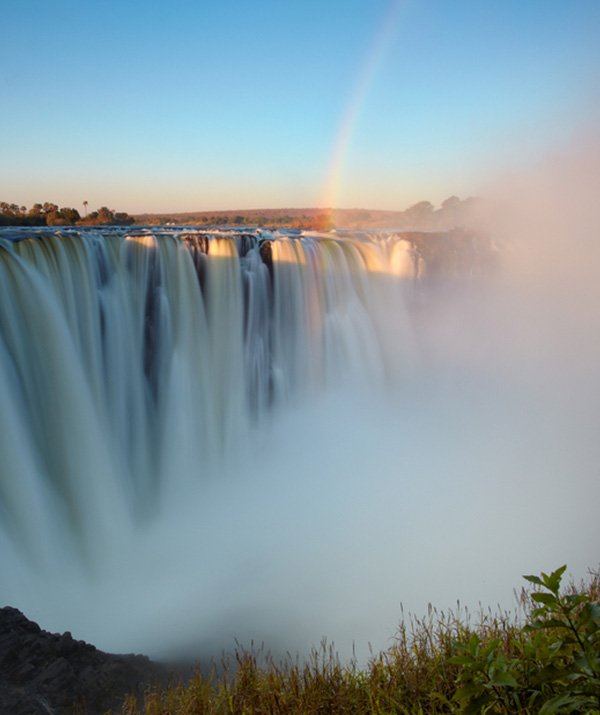 Victoria Falls, between Zambia and Zimbabwe, is a stunning sight at dawn. The rising sun bathes the rocky edge in golden light as the Zambezi River flows gracefully over, creating rainbows in the mist. It's a powerful and beautiful testament to nature's majesty.