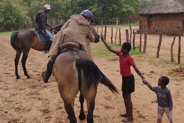 horse-riding-village-tour-local-children-victoria-falls-zimbabwe.jpg Tourists on horseback greeting local African children in traditional village near Victoria Falls Zimbabwe showcasing authentic cultural horse riding experience